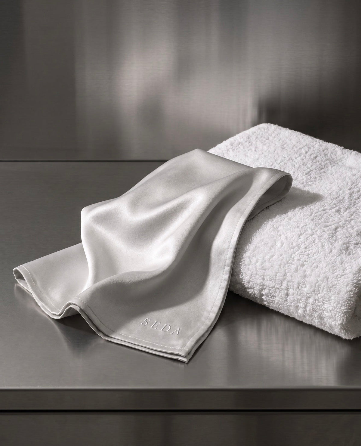 A smooth SEDA silk face cloth beside a folded cotton terry towel, showing the difference between a flat charmeuse weave and a raised looped pile